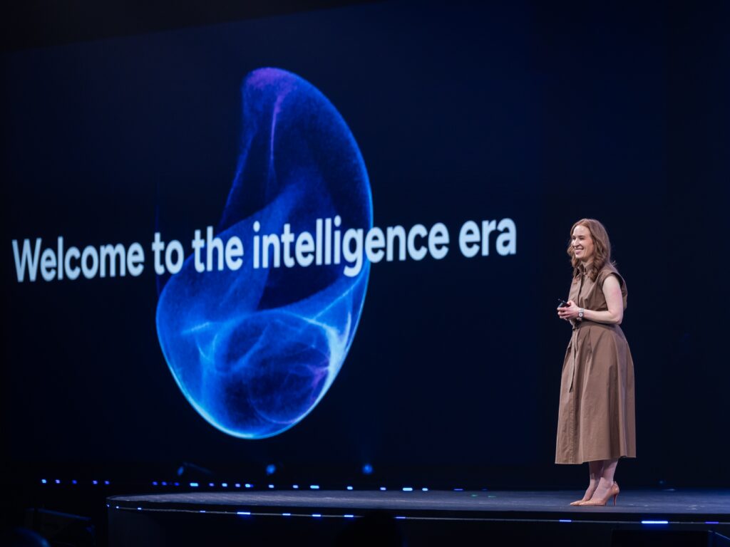 Kaseya CEO Rania Succar standing on stage giving a speech with a large screen to her left with a pixelated blue spherical shape on a black background with the words Welcome to the intelligence era across the center bold white letters. She is announcing the new Kaseya Intelligence platform.